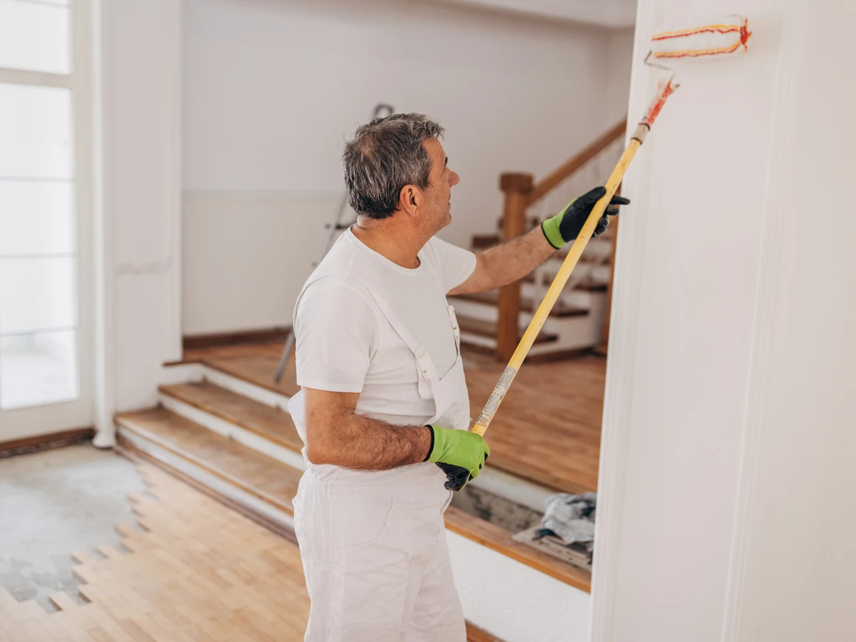 Decorator in white overalls rolling paint onto a wall beside a staircase