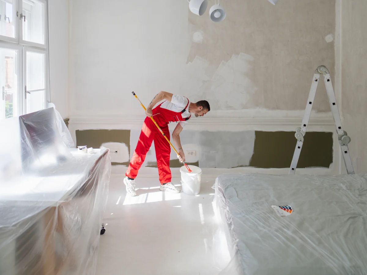 Painter in red overalls working in a room with the floor and furniture covered in plastic sheeting