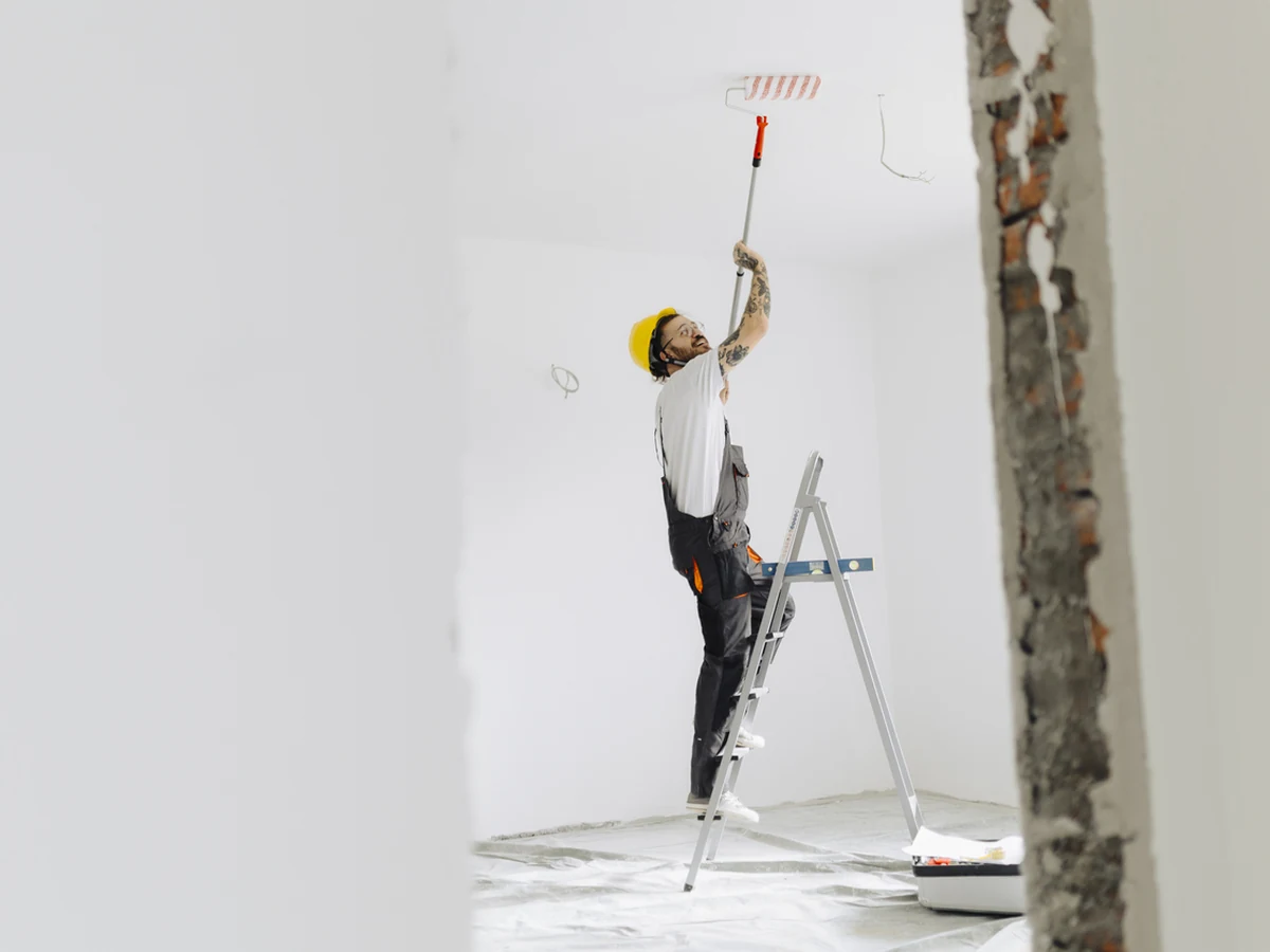 A decorator on a stepladder painting a high ceiling with a roller in a bright room